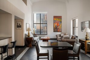 A modern living room with a sofa, chairs, a coffee table, and a TV. There are two windows with city views, and a poster on the wall. - The Manhattan Club (New York)