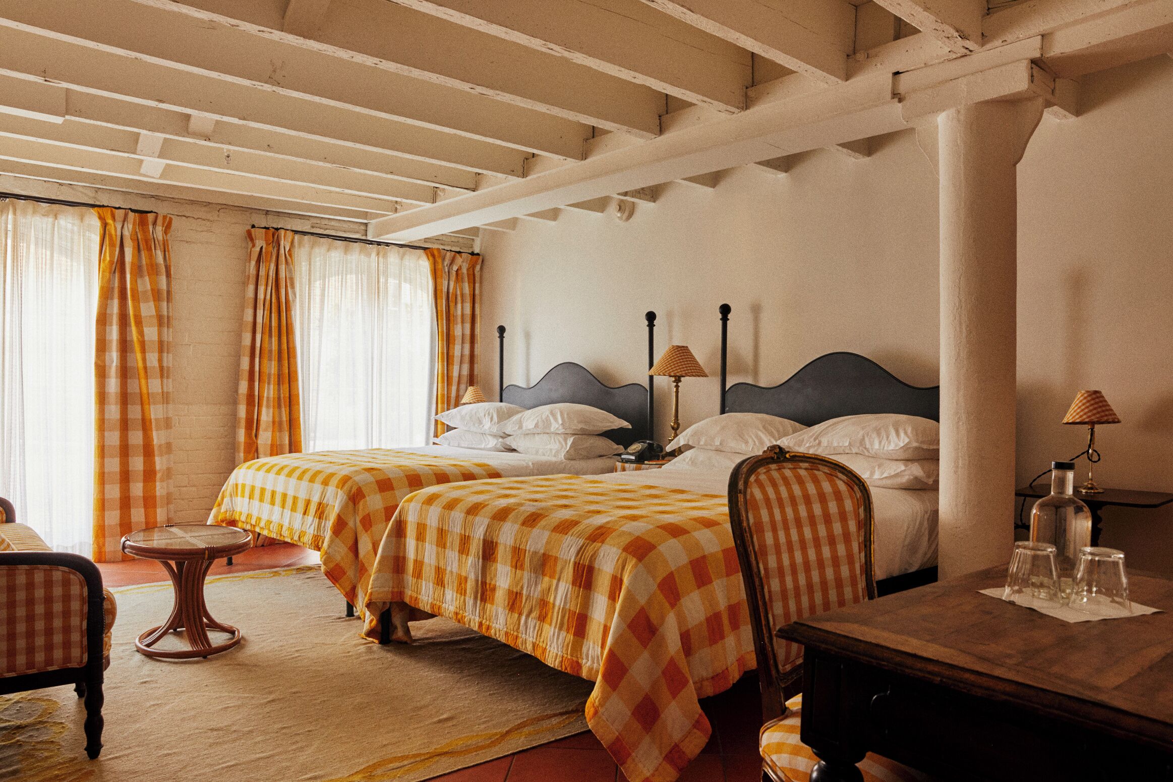 Quarto de hotel com duas camas, uma mesa redonda pequena, uma cadeira e uma escrivaninha de madeira com copos e uma garrafa.