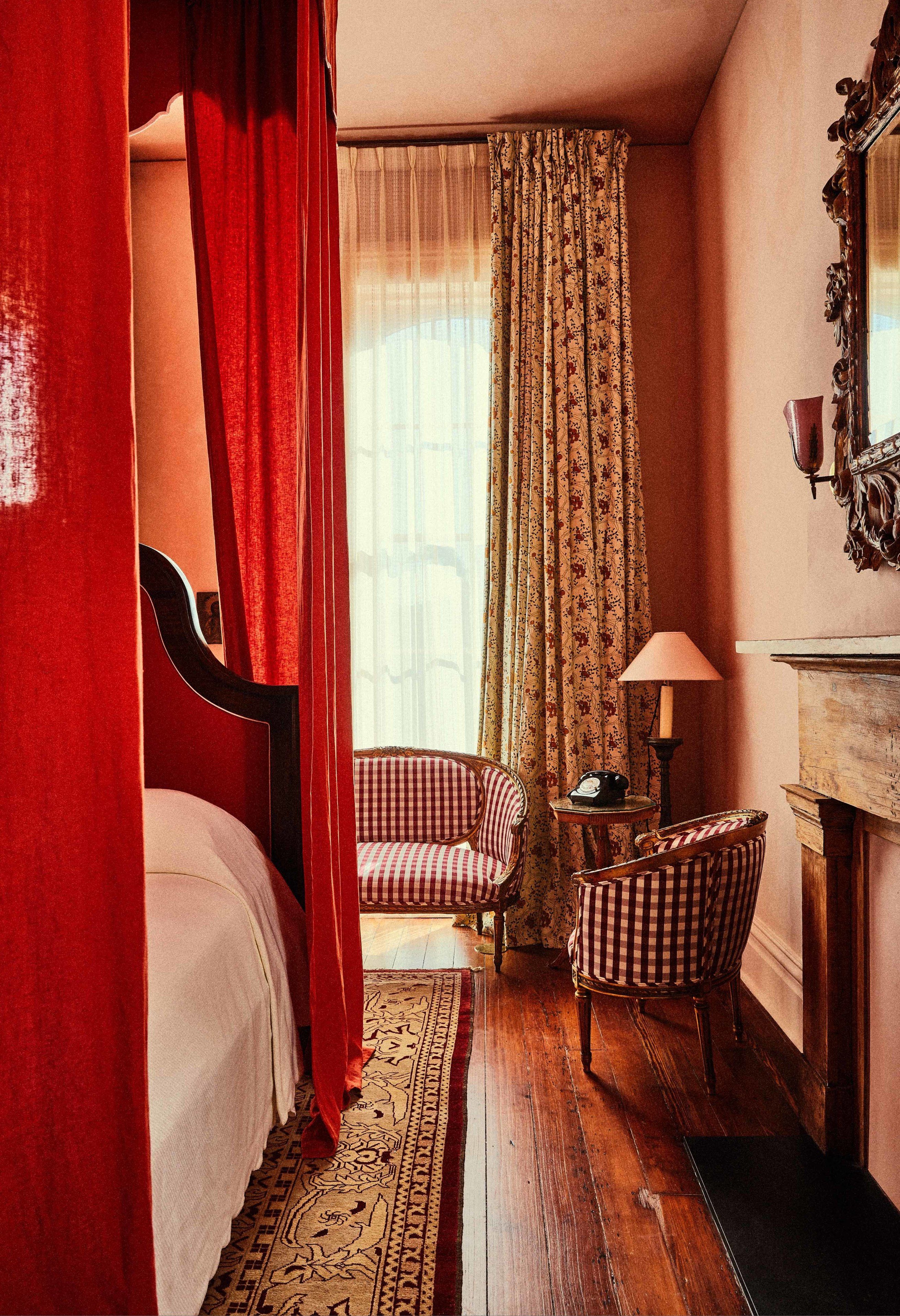 A bedroom with a four-poster bed, a checkered armchair, a small table with a lamp, and a wooden dresser.
