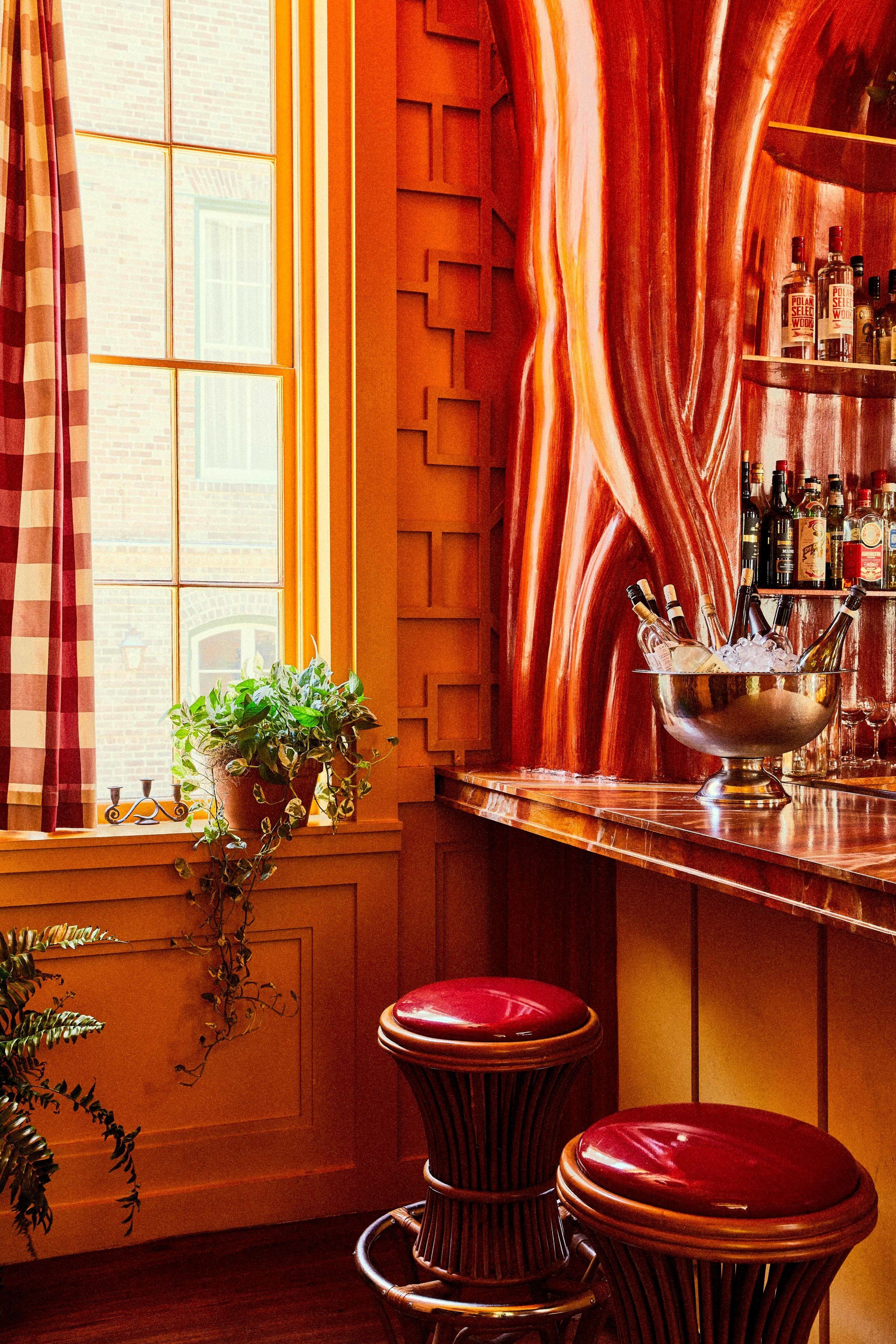 Una zona de bar con un mostrador de color cobre, un cubo metálico con botellas y dos taburetes rojos.