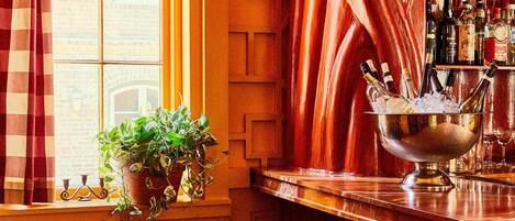 Una zona de bar con un mostrador de color cobre, un cubo metálico con botellas y dos taburetes rojos.