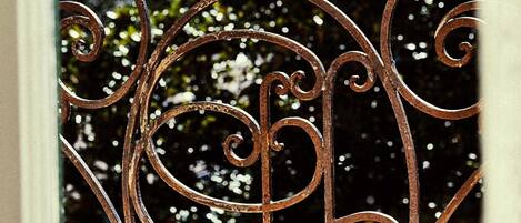 A wrought iron balcony railing with intricate designs, set against a backdrop of trees and a clear sky.