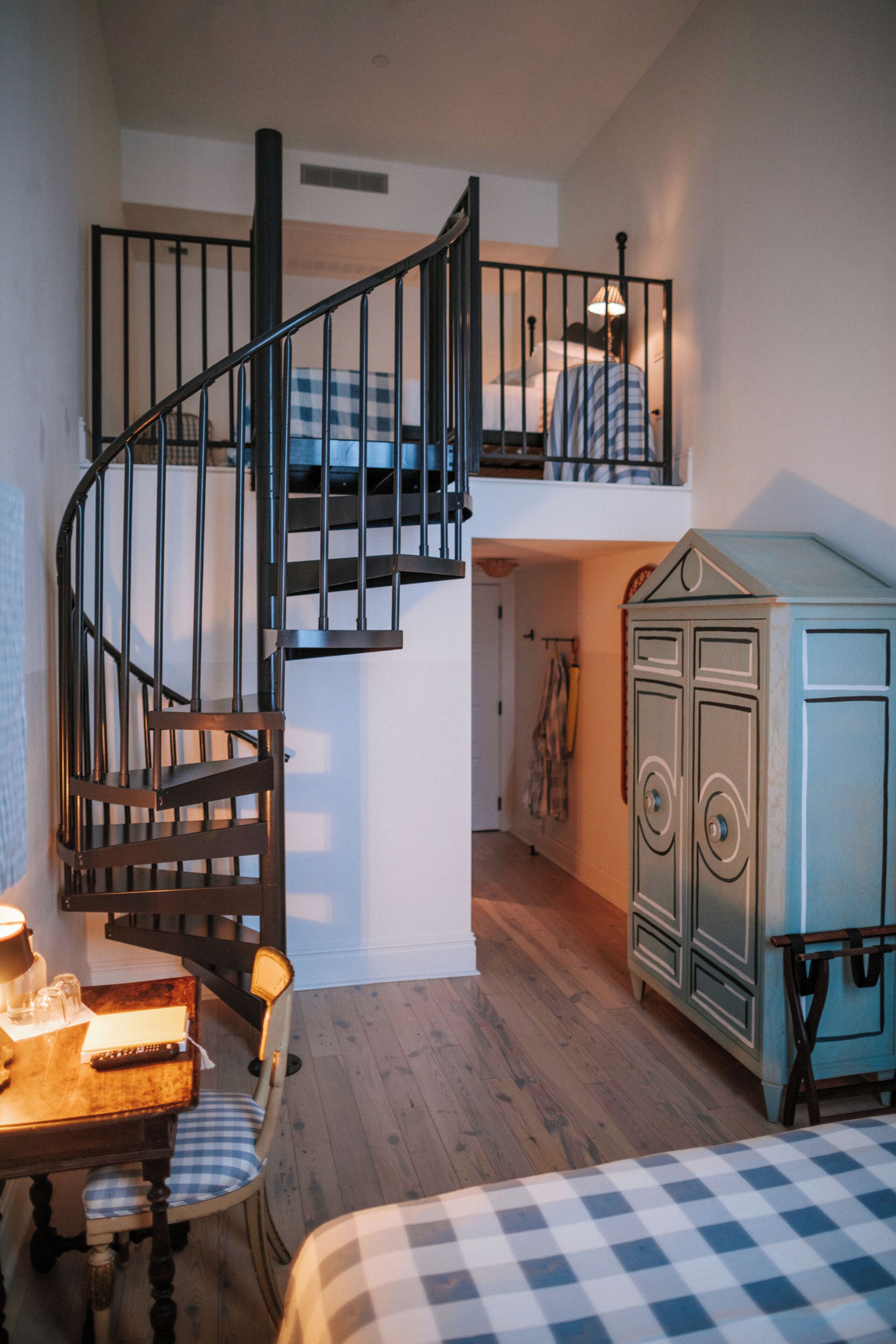 A bedroom with a checkered bedspread, a wooden chair, a small table with a lamp, a staircase with a railing, and a wardrobe.
