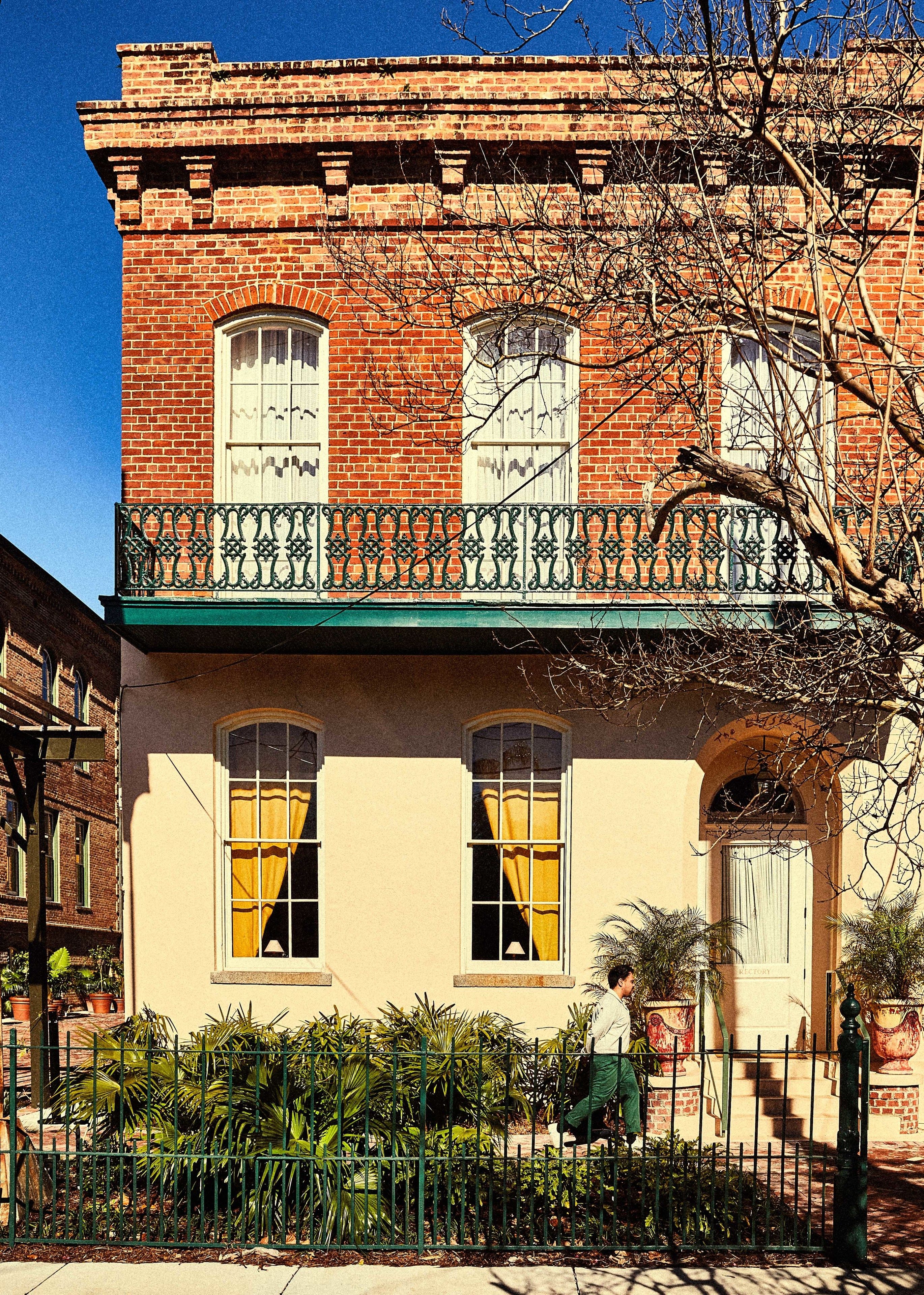 A two-story building with a brick facade, a balcony with ornate iron railings, and a gated entrance with potted plants.