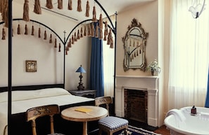 A bedroom with a four-poster bed, a fireplace, a mirror, and a small round table.
