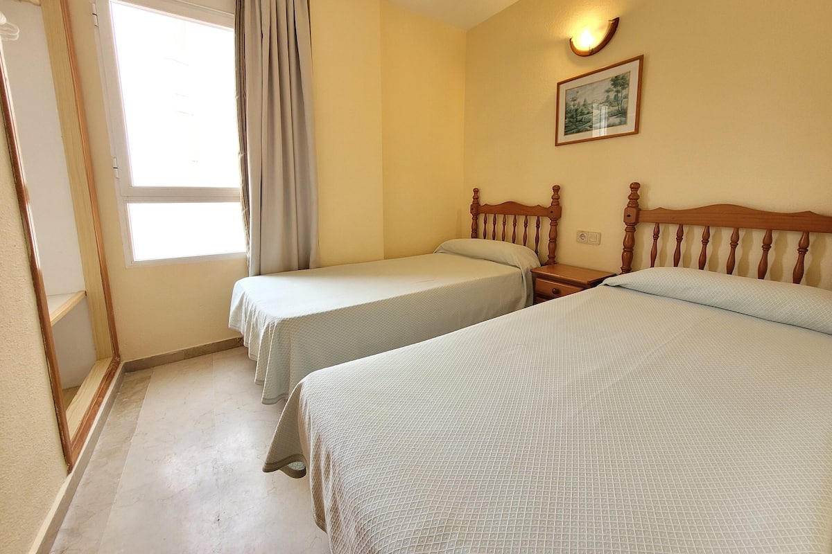 A hotel room with two single beds, a window with curtains, a wall-mounted light, and a framed picture.