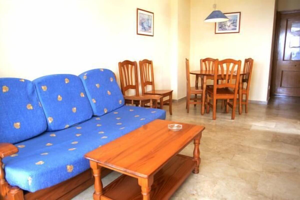 A living room with a blue sofa, wooden chairs, a wooden coffee table, and a dining table with chairs.