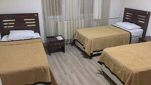 Une chambre d’hôtel avec trois lits simples, chacun recouvert d’une couverture marron et agrémenté d’oreillers blancs. Une petite table de chevet en bois se trouve entre les lits, sur laquelle reposent un téléphone et une petite lampe. La chambre est dotée d’un plancher en bois et d’une fenêtre ornée de rideaux légers.