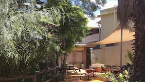 An outdoor patio area with tables and chairs, surrounded by lush greenery and a building with a balcony. - Hotel Cunnamulla (Cunnamulla)