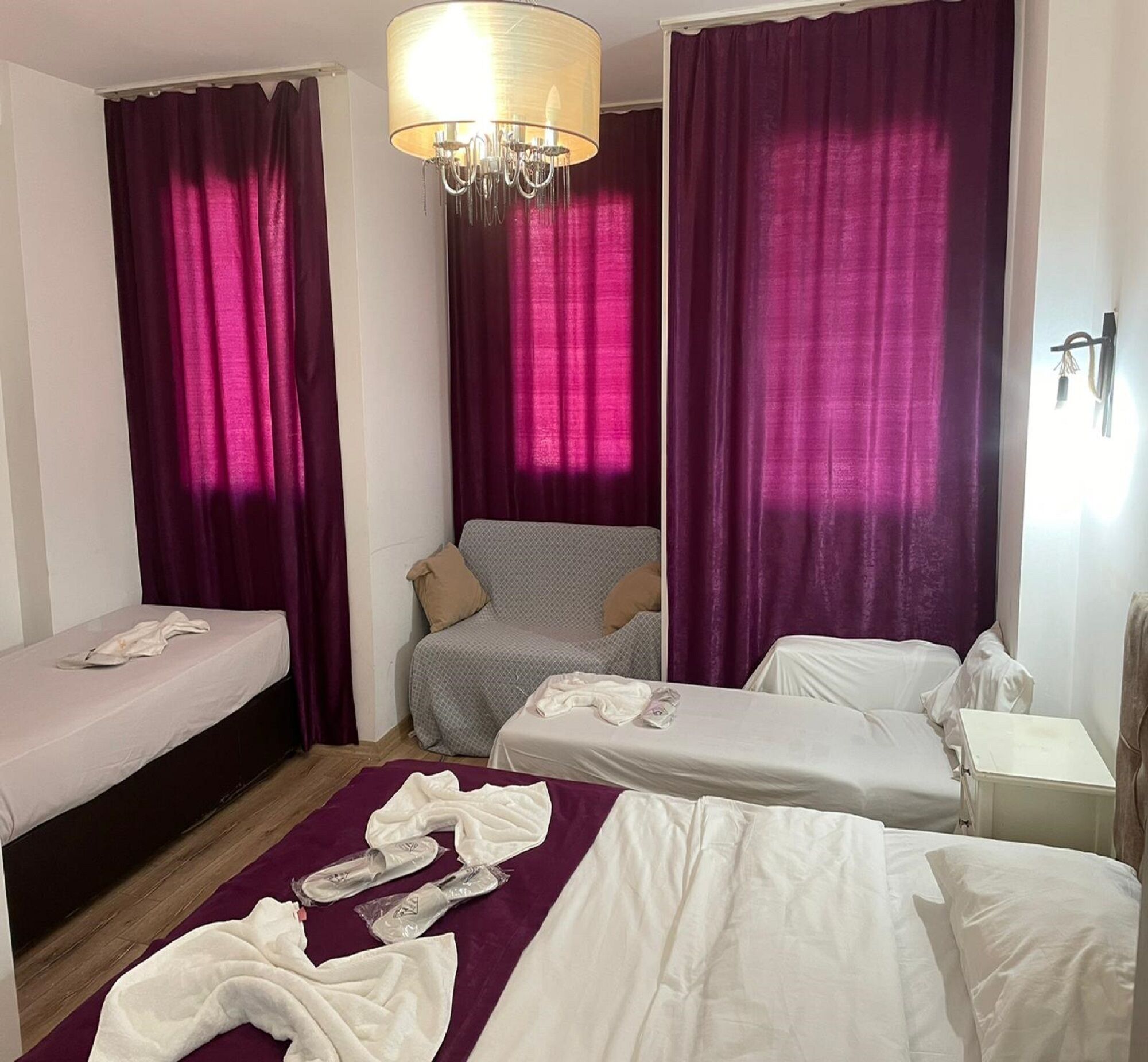 A hotel room with two single beds, a sofa, a chandelier, and purple curtains.