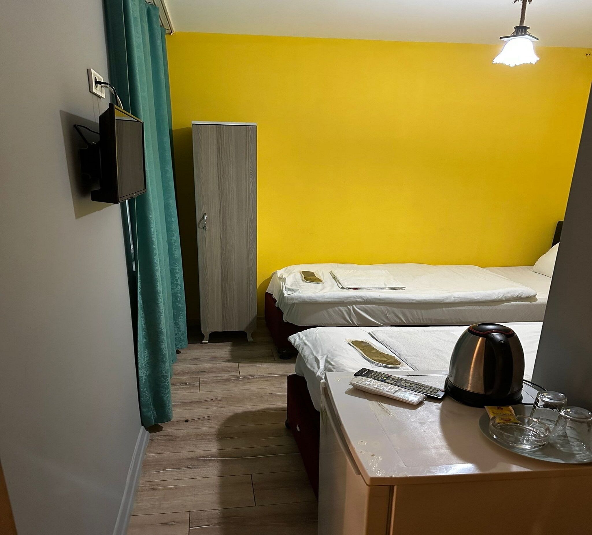 A small, brightly colored room with two beds, a wardrobe, a desk with a kettle, and a lamp.