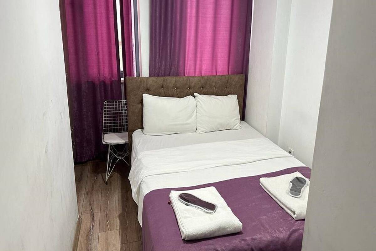 A small, neatly made bed with white linens and a purple bedspread, a white chair, and pink curtains in a compact room with wooden flooring.