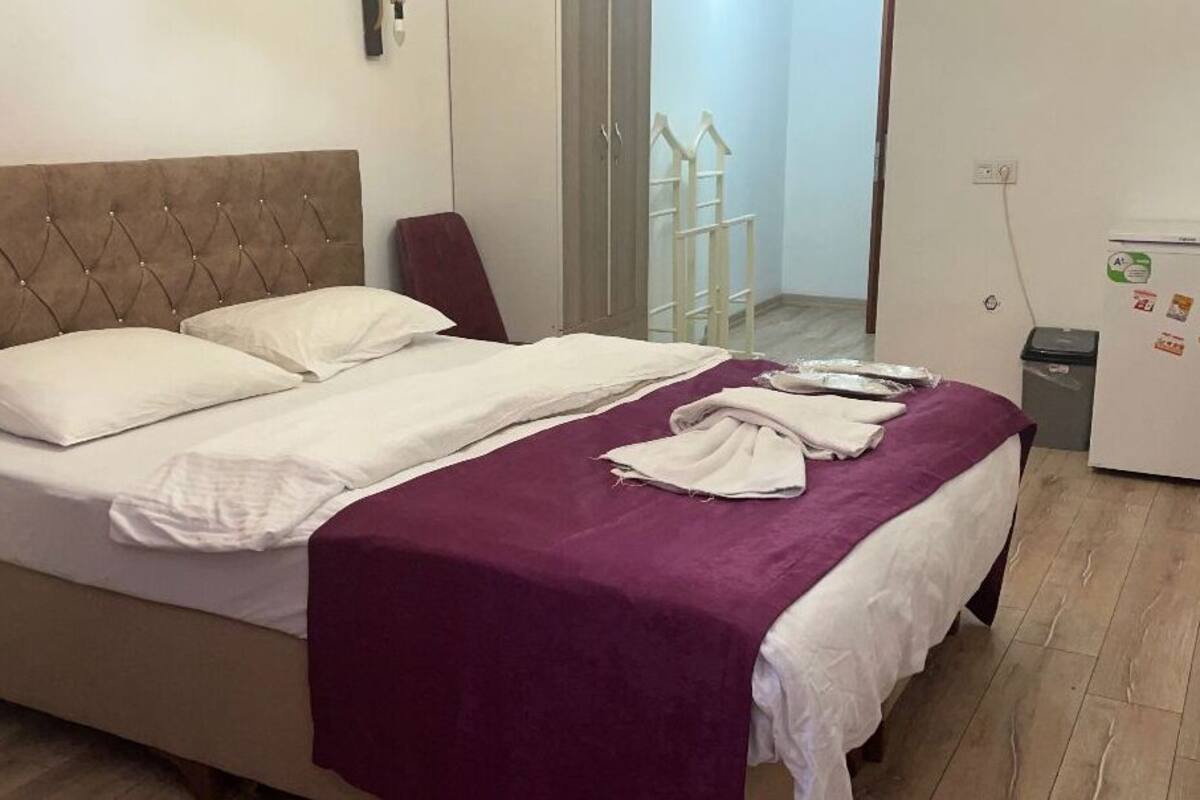 A bedroom with a bed, a wardrobe, a small refrigerator, and a trash bin.
