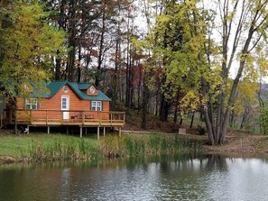 A wooden cabin with a green roof, situated by a calm lake, surrounded by trees with autumn foliage.