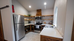A compact kitchen with a stainless steel refrigerator, a microwave, and a stove. There are wooden shelves above the counter, and a window on the right side. -  Both cottages rent as one unit,  Enjoy Privacy & Comfort, Very Family friendly (Branson)