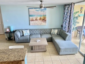 A living room with a grey sectional sofa, a wooden coffee table, a ceiling fan, and a beach-themed painting on the wall. - Lovely apartment with stunning ocean and marina views (Fajardo)