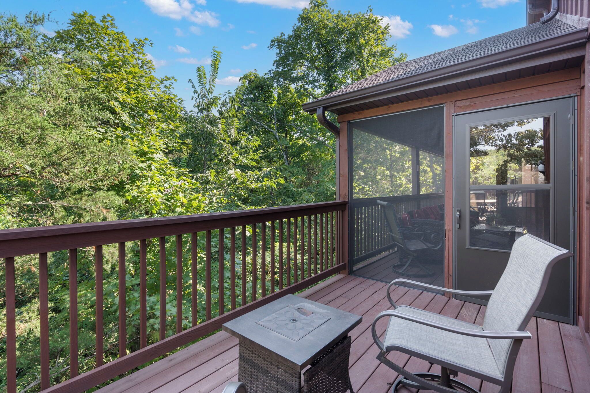 A wooden deck with a glass door leading to a covered outdoor area, surrounded by lush greenery.