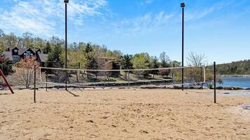 A sandy beach volleyball court with a net, surrounded by trees and a house in the background.