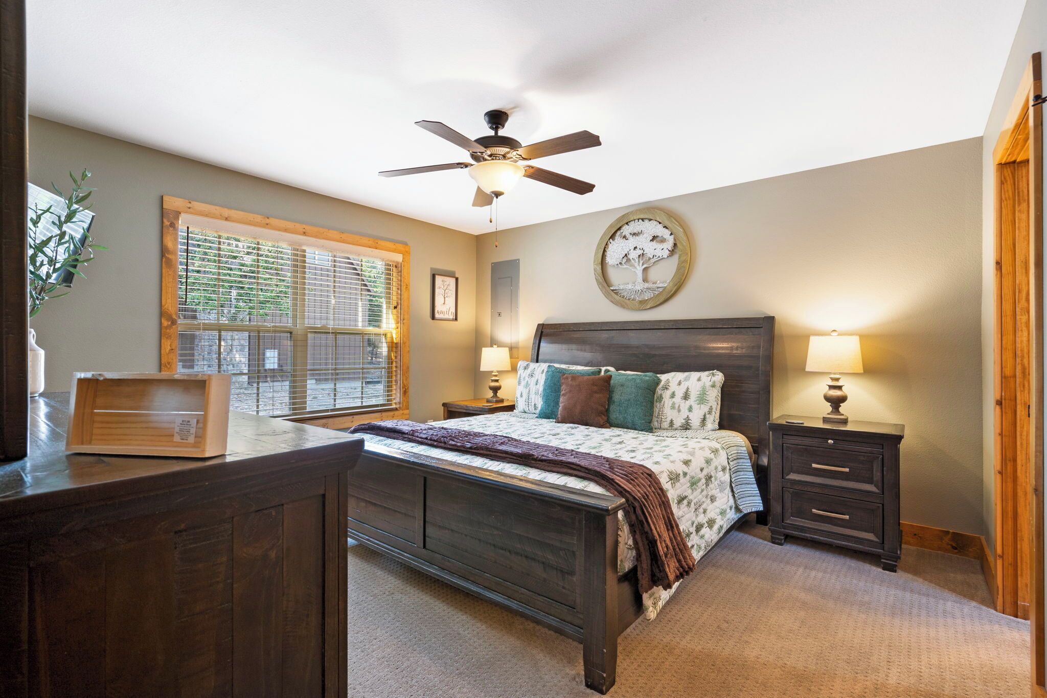 A bedroom with a bed, bedside tables, a ceiling fan, a window with blinds, and a wall-mounted clock.