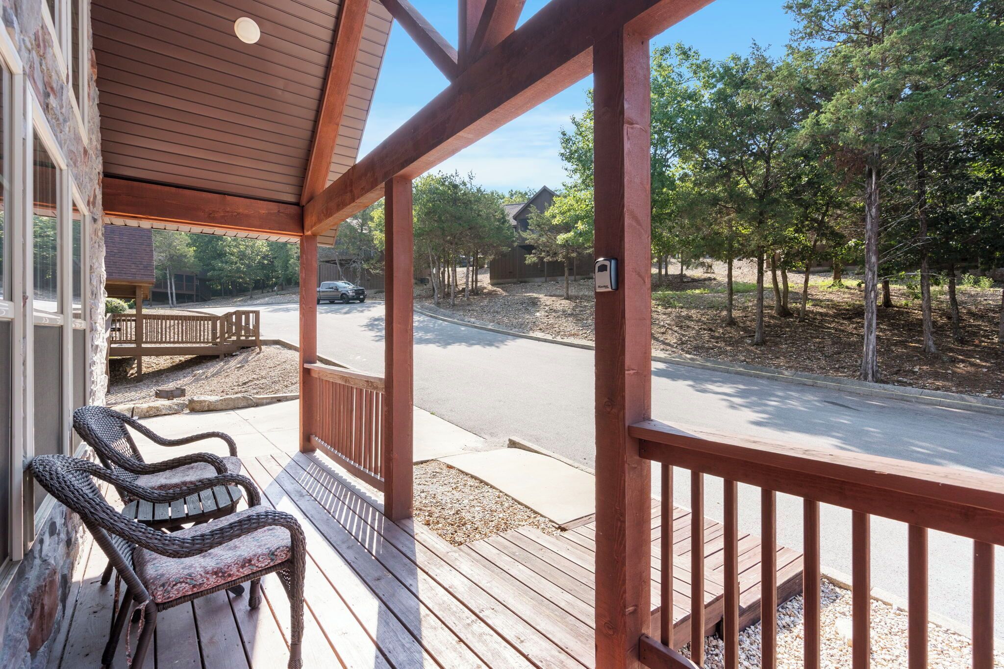 Een houten veranda met twee rieten stoelen, een brievenbus, en uitzicht op een straat met bomen en huizen.