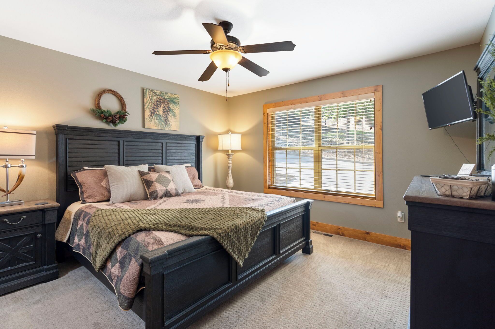 A bedroom with a dark wooden bed, a ceiling fan, a window with blinds, a TV on a stand, and a nightstand with a lamp.