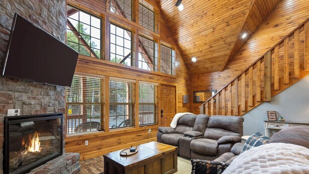 A cozy living room with a fireplace, large windows, and a wooden staircase. - Close to Outdoor Pool, Jetted Tub, Rustic Lodge! (Reeds Spring)