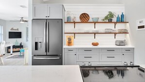 A modern kitchen with a stainless steel refrigerator, white cabinetry, and a black stove top. There are wooden shelves above the counter holding dishes and a potted plant. - Beachcomber Bungalow: Slow Down, East of A1A, Heated Pool, Hot Tub & Beach Gear (Vero Beach)