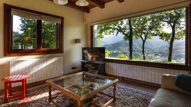 Un salon chaleureux avec une grande fenêtre offrant une vue panoramique sur les montagnes, une table basse en verre, un fauteuil rouge et un canapé.