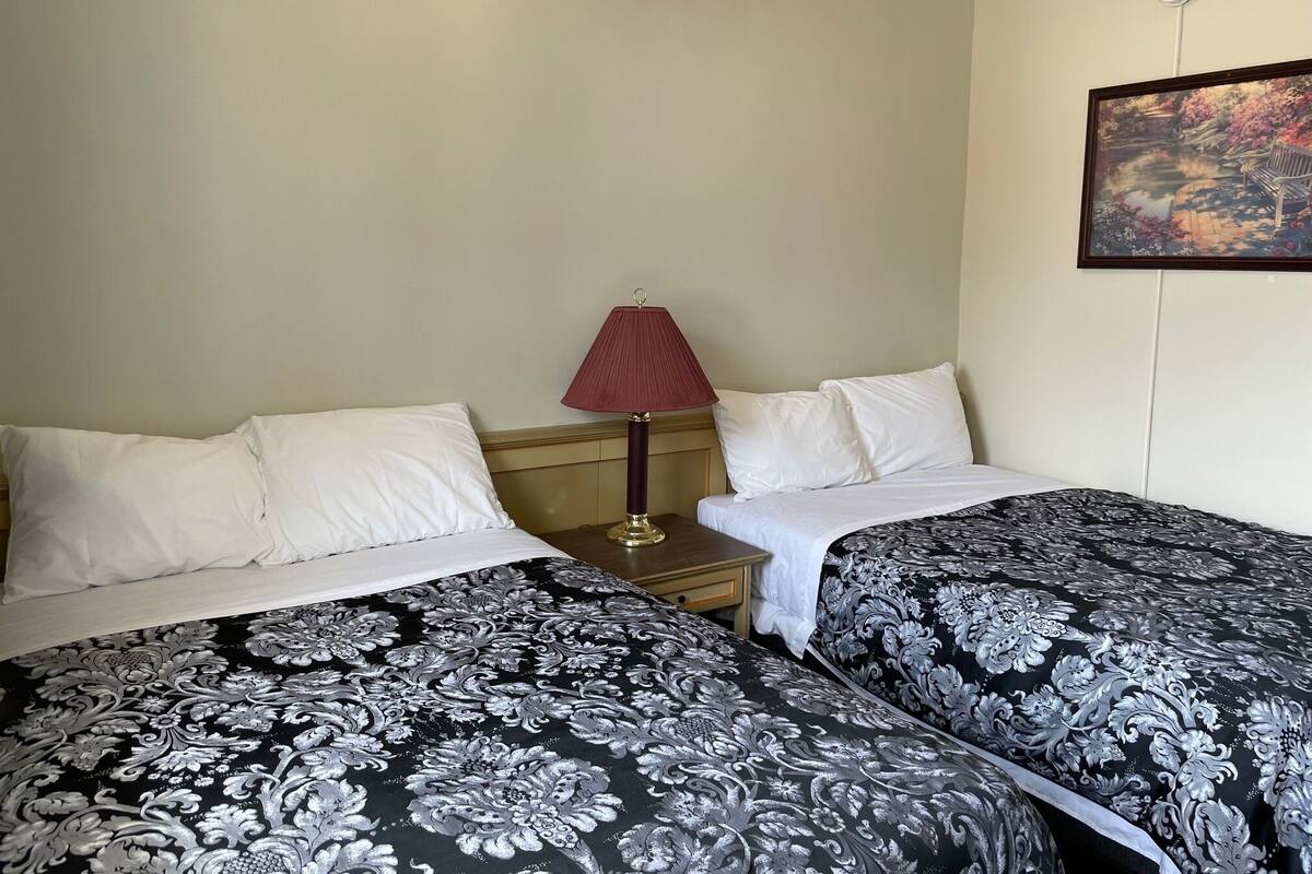 A hotel room with two beds, each with a black and white patterned bedspread, a wooden nightstand with a red lamp, and a painting on the wall.