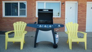 A barbecue grill with two yellow Adirondack chairs in front, set against a brick building with numbered doors.
