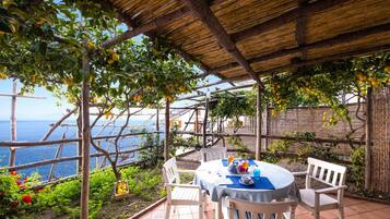 A covered outdoor dining area with a table and chairs, surrounded by lemon trees and a view of the sea.