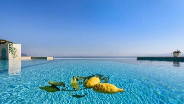 A clear blue pool with a view of the sea, a concrete structure with greenery, and a potted plant on the right.