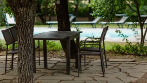 An outdoor seating area with a table and chairs, surrounded by trees and a pool in the background.