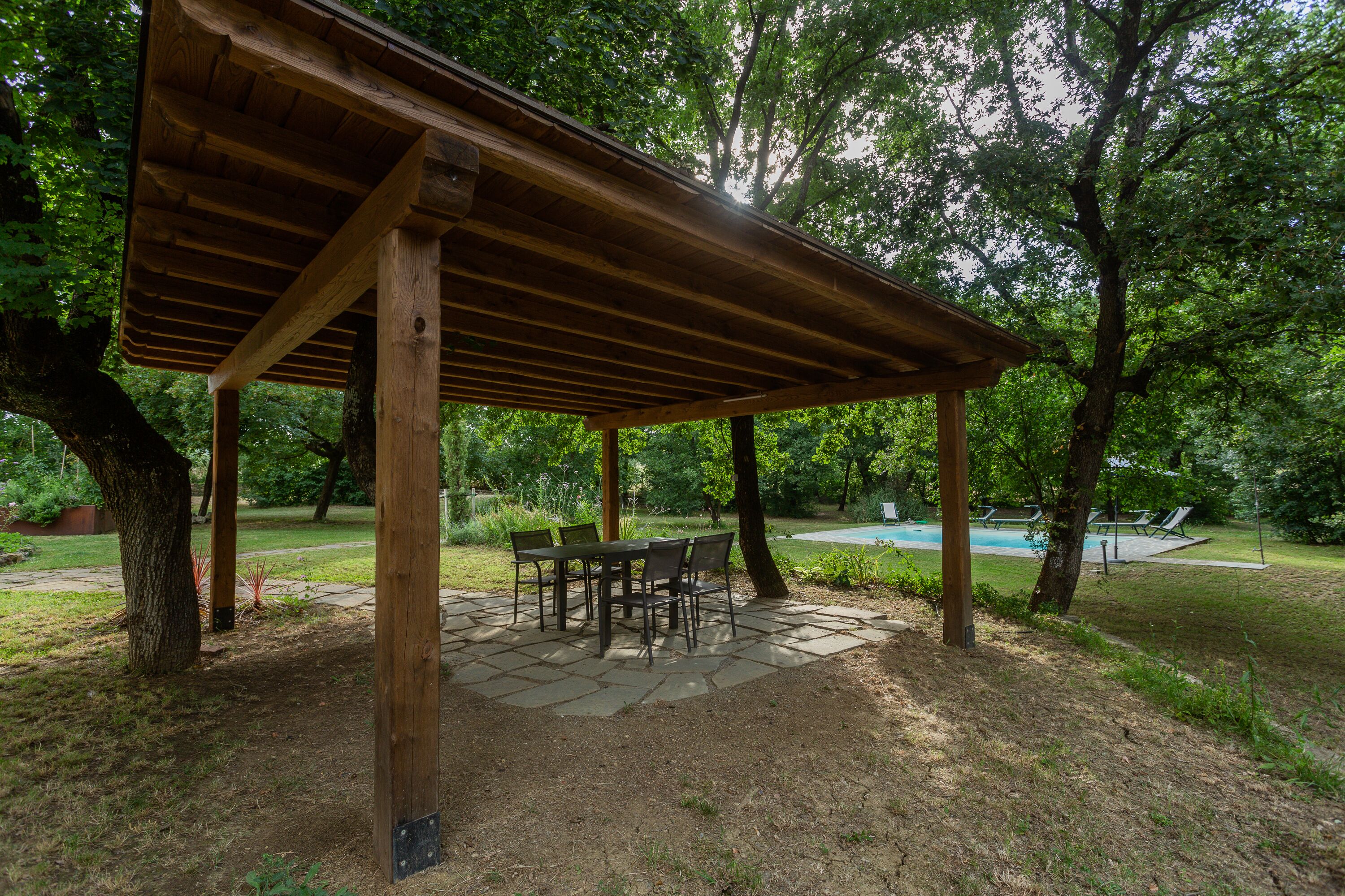 A wooden pergola with a stone patio table and chairs, surrounded by trees and a swimming pool in the background.