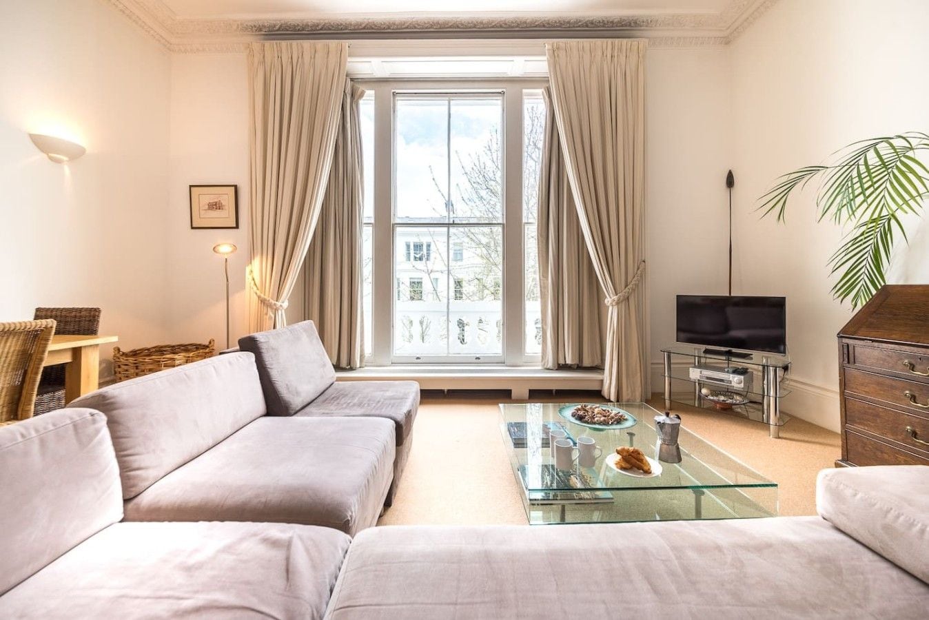 A modern living room with a sofa, a glass coffee table, a television, and a large window with curtains.