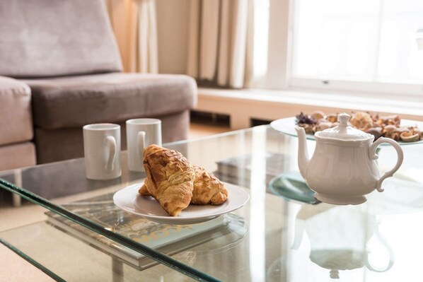 A glass-top table with a teapot and a plate of pastries, a sofa, and two mugs in a living room.