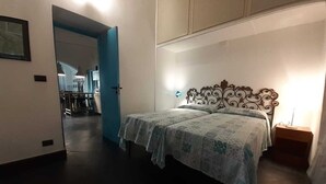 Une chambre à coucher avec un lit, une table de chevet et une lampe fixée au mur. Une porte donne accès à une autre pièce.