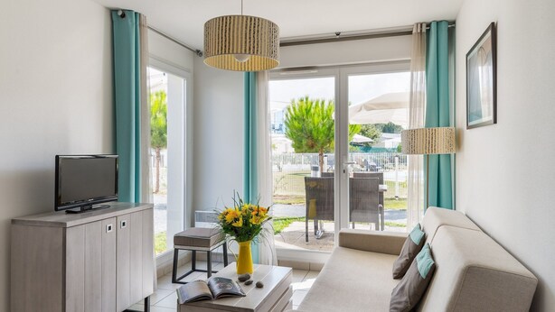 Ein modernes Wohnzimmer mit einer Couch, einem Fernseher auf einem Schrank, einem kleinen Tisch mit Blumen und Blick auf die Terrassenanlage.