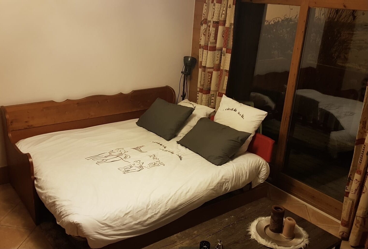 Ein Bett mit weißer Bettdecke und zwei Kissen, ein hölzernes Kopfende, ein Nachttisch mit Kerze und ein Fenster mit Vorhängen.