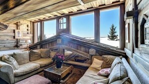 Een houten blokhut van binnen, met een gezellige zithoek, grote ramen en uitzicht op het besneeuwde landschap.