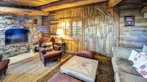 Een landelijk chaletinterieur met een stenen openhaard, houten wanden, een gezellige zithoek met een comfortabele fauteuil en bank, en een kleine tafel met een lamp.