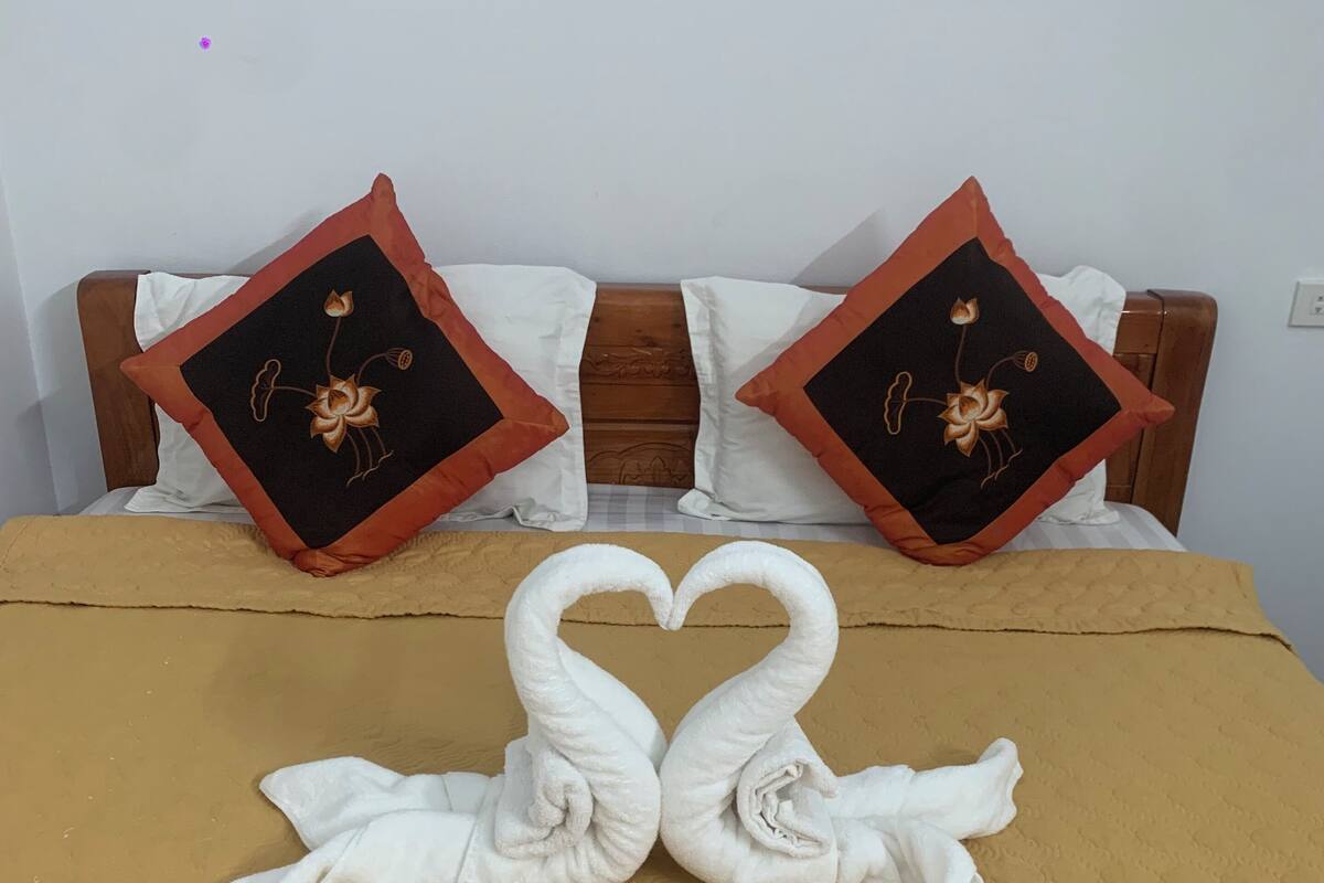 Una cama con una colcha marrón, dos almohadas con diseños florales y dos toallas dispuestas en forma de corazón.