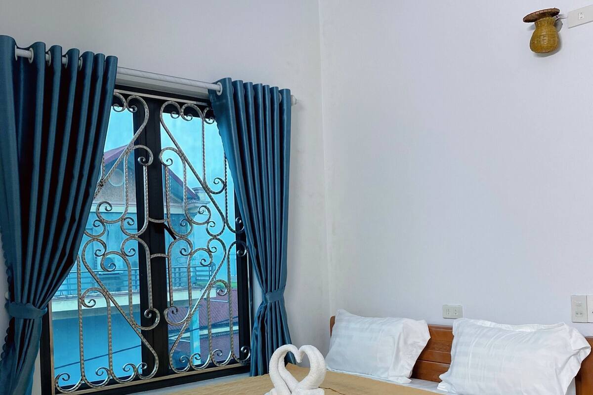 Habitación de hotel con cama, almohadas blancas, cabecero de madera y vistas a través de una ventana con cortinas azules.