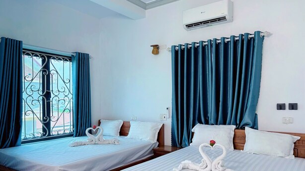 A hotel room with two beds, a window with decorative iron bars, blue curtains, an air conditioning unit, and a thermostat on the wall. - Tamcoc White Swan Hostel (Nam Hoa Lu)