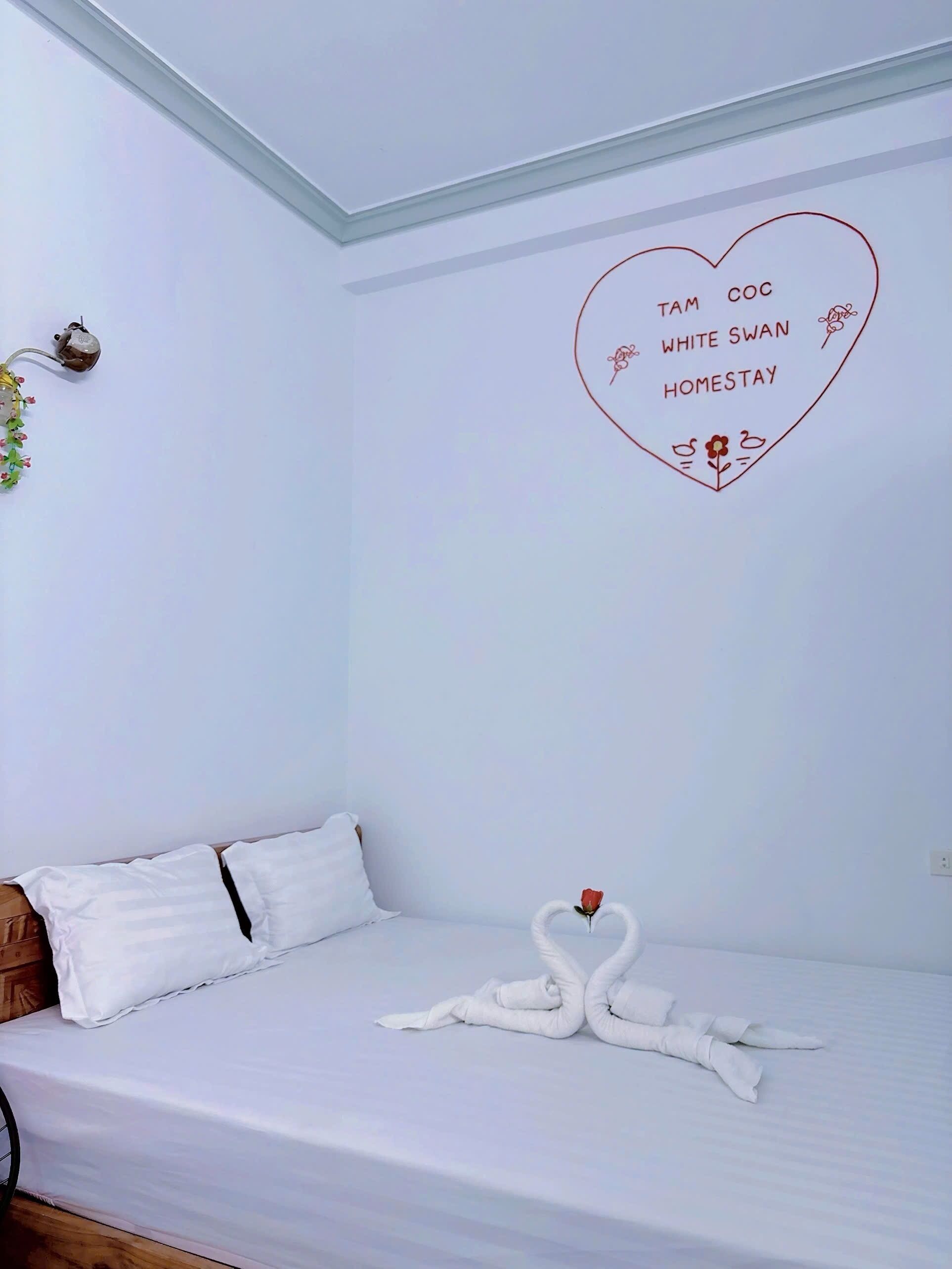 A hotel room with a bed, white bedding, a heart-shaped decoration on the wall, and a swan towel arrangement.