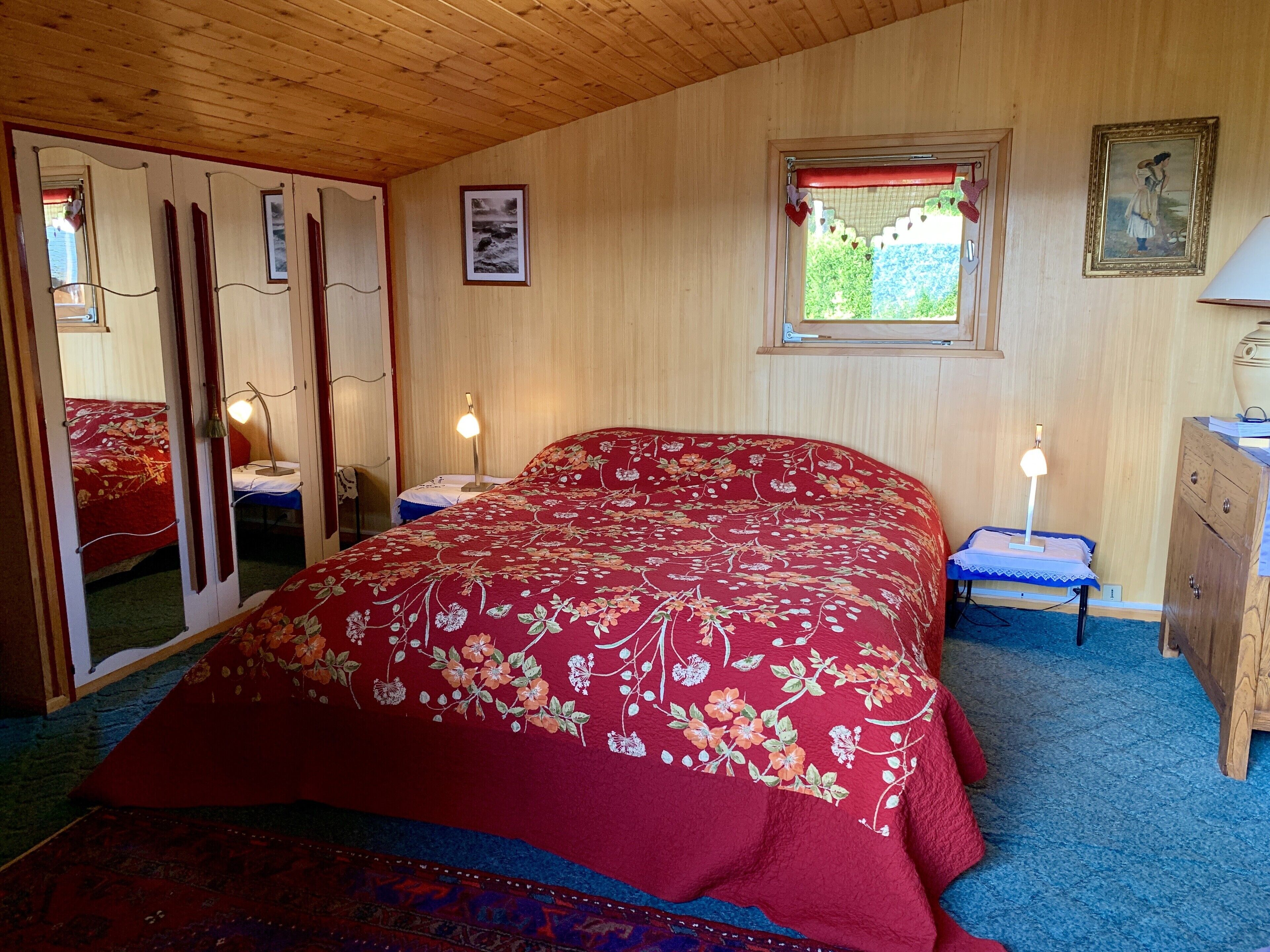 Un dormitorio con techo de madera, una cama con colcha floral, un aparador, una mesita de noche con lámpara, una ventana con vistas a vegetación y un espejo con colcha roja.