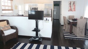 Ein Wohnzimmer mit Fernseher auf einem Schrank, ein Essbereich mit Stühlen, eine Küche mit Geräten und einem Deckenventilator.