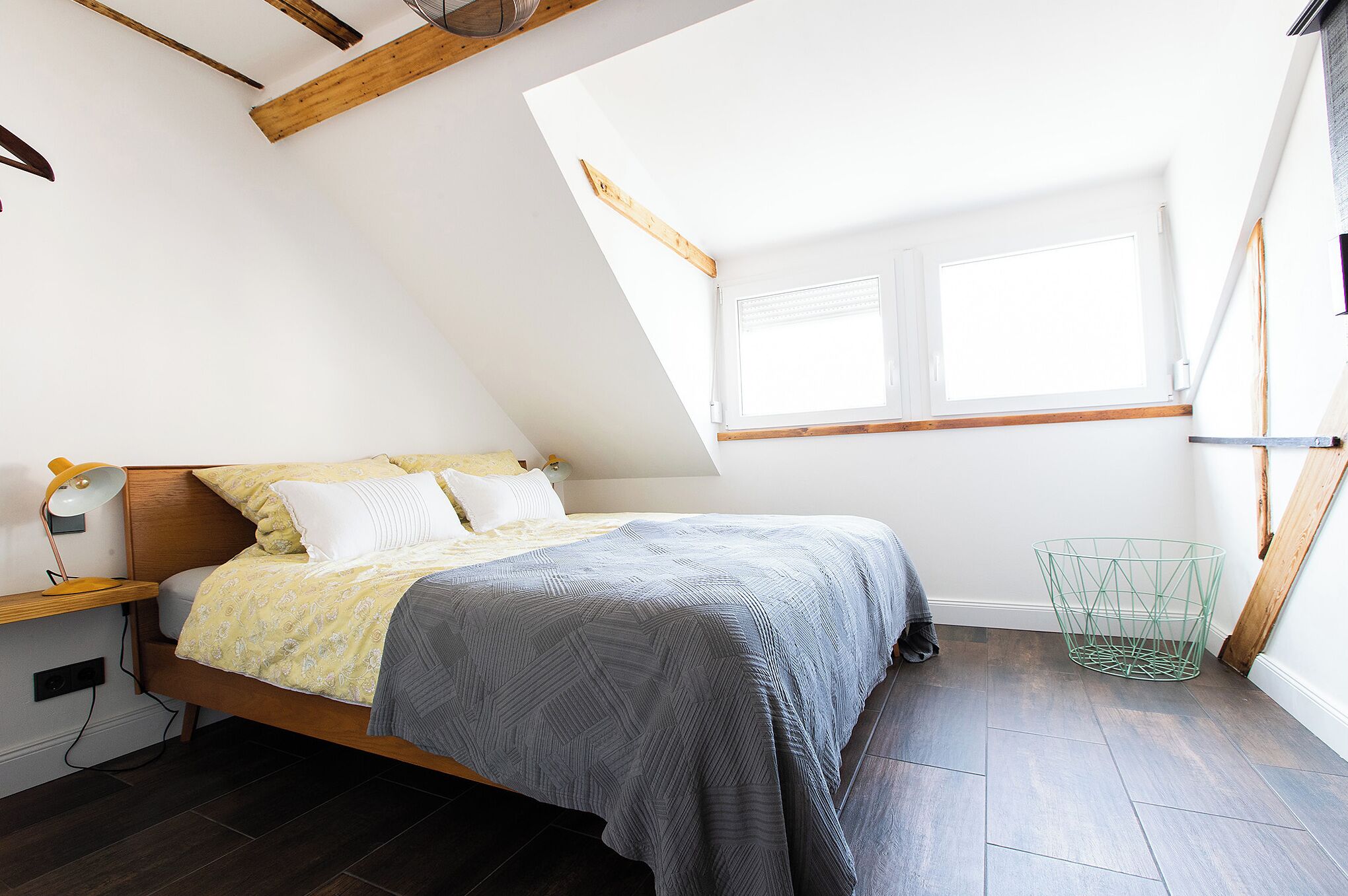 Une chambre avec un plafond en pente, un lit recouvert d’une couvre-lit à motifs, une table de chevet avec une lampe et une fenêtre avec des stores.
