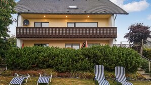 Une maison à deux étages avec un balcon, un petit jardin et deux fauteuils de jardin à rayures.