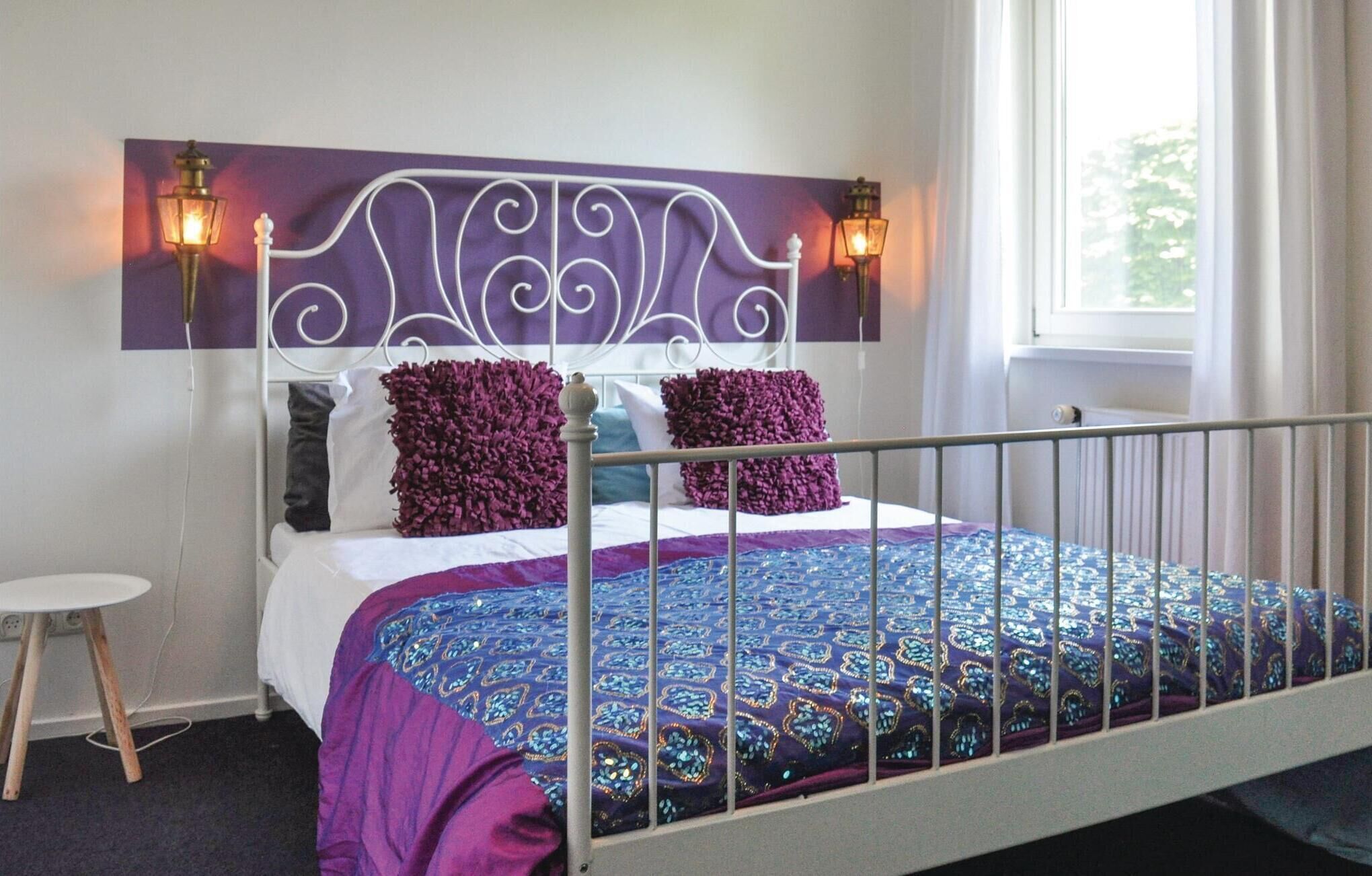 A neatly made bed with a purple and blue patterned bedspread, a white metal headboard with decorative lights, and a small round table beside the bed.
